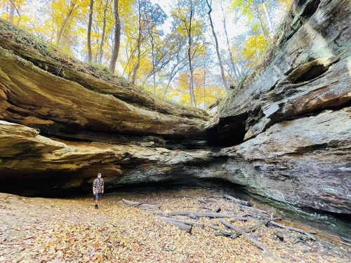 A person stands in a rocky cave surrounded by autumn trees and fallen leaves, with sunlight filtering through.