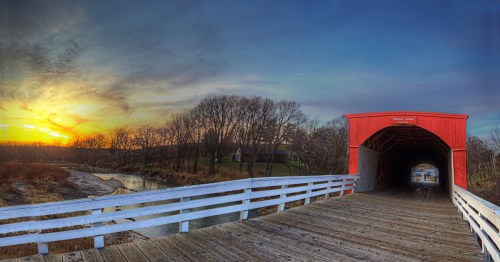 A scenic view of a red covered bridge at sunset, with a river and trees in the background.
