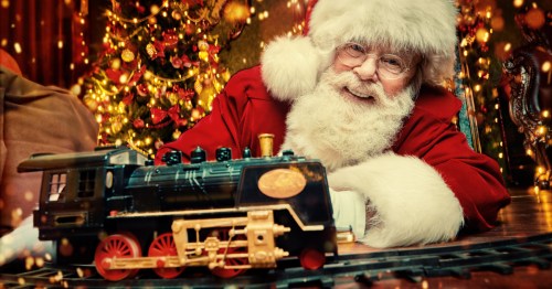 Santa holding a model train car in front of a Christmas Tree.