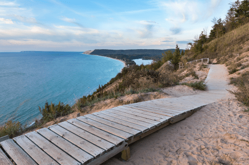A wooden boardwalk winds through sandy terrain, overlooking a scenic coastline and calm blue waters under a cloudy sky.