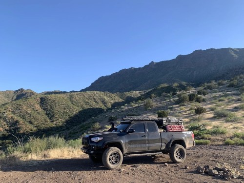 A rugged truck parked on a dirt road with mountains and greenery in the background under a clear blue sky.