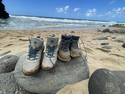 Two pairs of muddy hiking boots resting on a rock by a sandy beach with waves and a blue sky in the background.