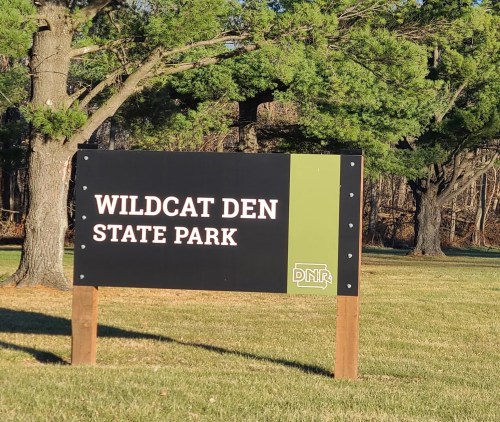 Sign for Wildcat Den State Park, featuring green and black colors, surrounded by trees and grassy area.