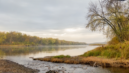 A calm river scene with trees along the shore, under a cloudy sky, reflecting autumn colors in the water.