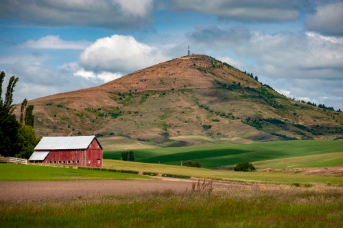 A red barn sits in a green field with a hill in the background, topped by a communication tower under a cloudy sky.