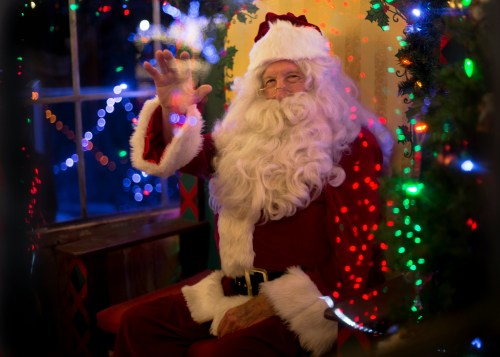 A cheerful Santa Claus waves from a cozy, decorated room filled with colorful Christmas lights.
