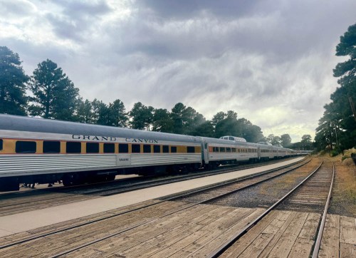 A vintage train labeled "Grand Canyon" parked on a wooden platform surrounded by trees under a cloudy sky.