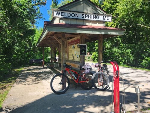 Bicycles parked at a rustic train station in Weldon Spring, MO, surrounded by lush greenery and a clear blue sky.