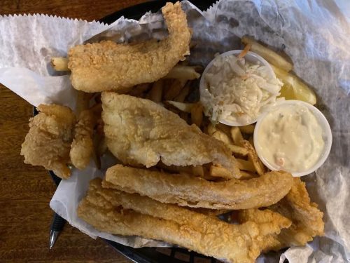 A basket of fried fish fillets, French fries, coleslaw, and dipping sauce on a bed of parchment paper.