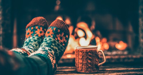 A person with wool socks sits with their feet near a fireplace with a mug.