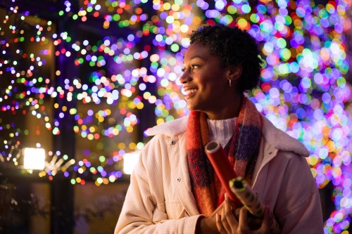 A smiling woman holds gift wrap, standing in front of colorful holiday lights.