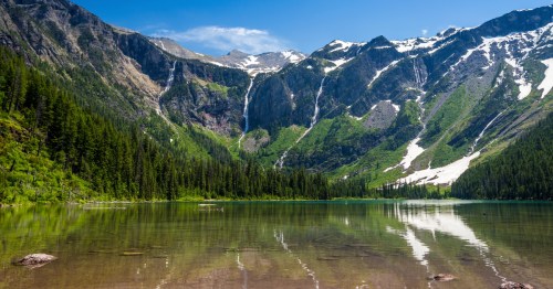 An alpine lake in Glacier National Park surrounded by bold mountains, waterfalls, and green trees.