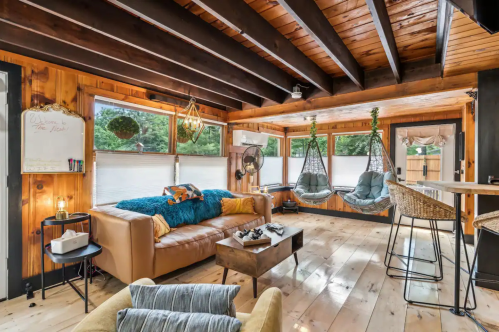 The living area of The NEST Haven Airbnb property in Newfield, Maine. The room has a chair, couch, and two swing chairs. It is rustically decorated.