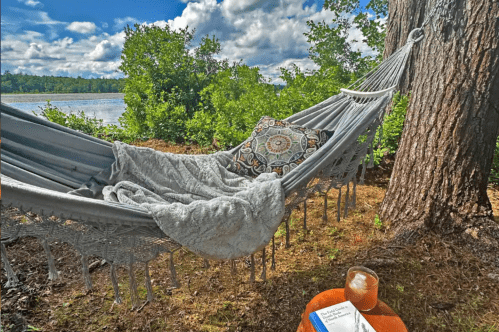 A hammock with a pillow and blanket hangs from on a tree at The Retreat at Eaglesville Airbnb in Athol, MA. A table with a chair and a drink is next to the hammock. A pond is in the background.