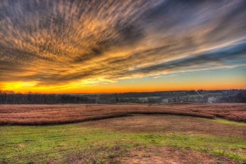 A cut-down farm field with forest in the distance under a cloudy sky with a setting sun casting it in shades of orange and purple.