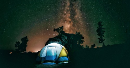 A glowing tent sits under a stunning starry sky with the Milky Way visible above silhouetted trees.