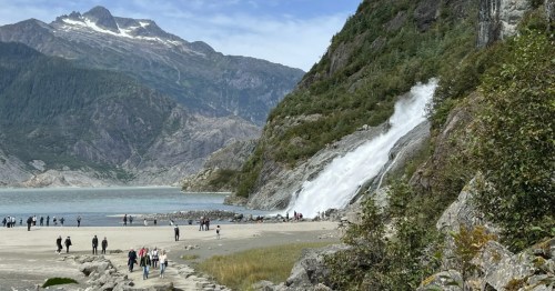 Nugget Falls cascades into Mendenhall Lake with hikers on the shore and mountains.