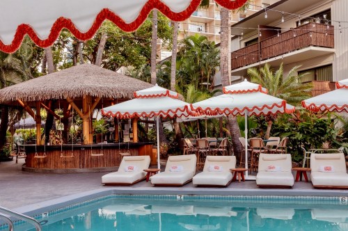 Retro pool deck at White Sands Hotel with white lounge chairs, red-trimmed umbrellas, and a thatched-roof swim-up bar.