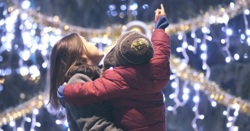 A mom holds a child in her arms as they both look up at twinkling holiday lights strung overhead.
