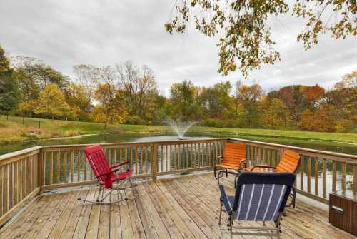 A deck with four chairs sits before a lake with a fountain, framed by an autumnal Iowa forest.