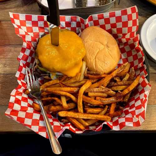 A large burger topped with cheese, with a knife stick inside it, accompanied by a pile of sweet potato fries in a basket.