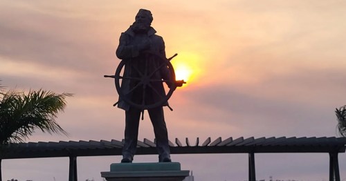 A statue of a ship captain holding the steering wheel of a ship with the sun setting in the distance.