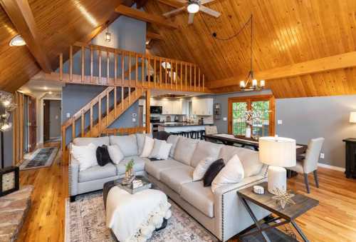 A cozy couch sits in the center of a cabin room with high ceilings and before a staircase leading to a loft.