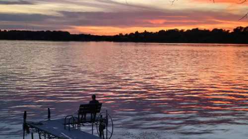 A dock stretches out onto a lake at dusk, with the waters reflecting a pink sky and framed by dark woods.