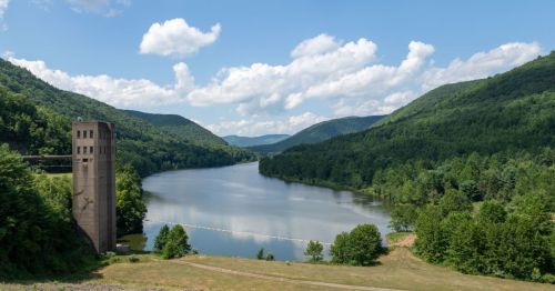 A panoramic view of a serene lake in Austin, Pennsylvania, surrounded by forested mountains, and a concrete structure is visible near the water's edge.