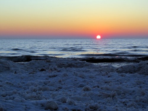 A snowy beach sits before open water and a sun setting into the horizon.