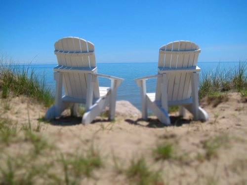 Two white Adirondack-style chairs sit on a sandy beach before open water under a blue sky.