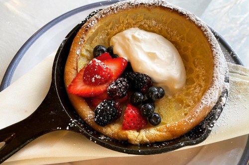A cast iron pan holding a dutch baby pancake topped with berries and whipped cream and sprinkled with powdered sugar.