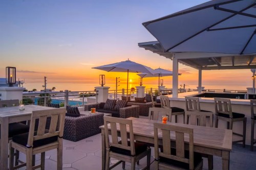 A rooftop dining experience with the vibrant orange colors of the lit-up sky as the sun sets on behind the beach horizon.