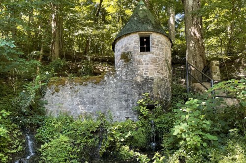 A rounded stone tower stands amidst greenery with a staircase on one side and water flowing from the rock underneath it.