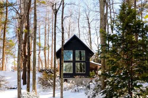 A cabin with tall picture windows overlooking a mountainous forest after a fresh layer of snow.