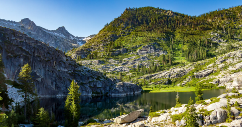 Beneath a blue sky, a white an red tent is set up beside a tranquil alpine lake.