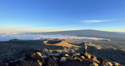 A hiker stands on a rugged outcrop, overlooking an arid mountain landscape.