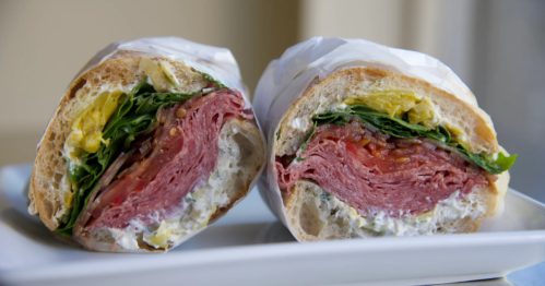 A crosscut of a delicious sandwich with artichoke cream cheese, arugula, red onion, pickled cucumber, balsamic reduction, pepperoncini, and tomato.