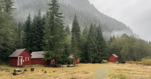 Beneath a misty, tree-covered mountain, derelict building with fading red paint are scattered in a field.