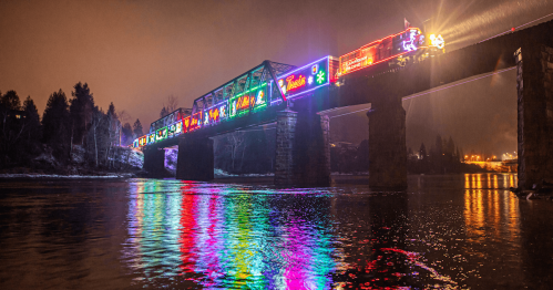 A colorfully lighted train cross a rail bridge over a river at night, its lights reflecting in the water.
