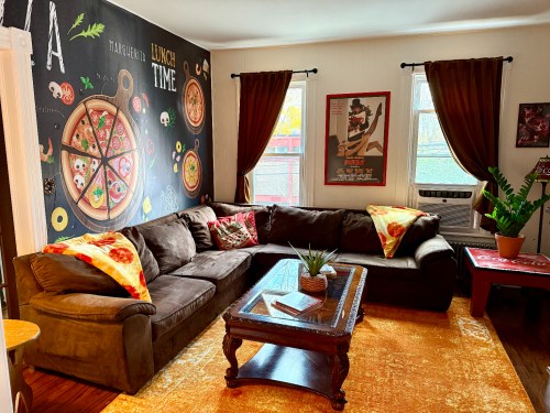 The living room at the "CT Pizza Palace" Airbnb in New Haven, CT. The pizza-themed room has a pizza mural, a brown couch with blankets resembling pizza slices, and other pizza-themed decorations.