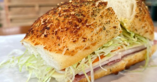 A sandwich with lettuce, meat, cheese, and tomato is made with a fresh bread roll and cut in half.
