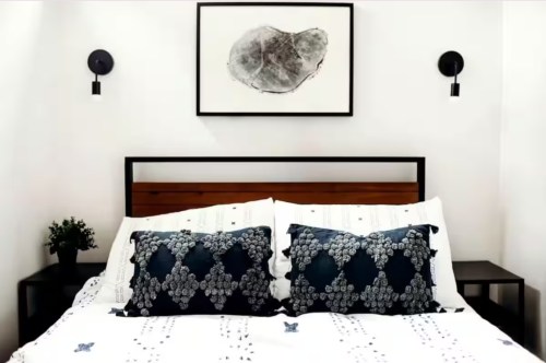 A cozy bed with a white comforter and black pillows under a framed abstract painting in a black and white room.