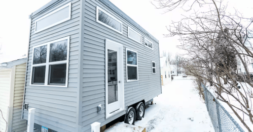 A bluish-grey tiny home with tons of window sits on a snow-covered property in Cedar Rapids, Iowa. There are bare, ice-covered trees to the right and a small cream-colored shed to the left. The sky and ground are the same shade of white.