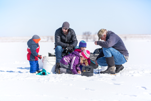 A family is ice fishing on a sunny, snowy day. A man and an older man are with three children bundled in winter gear.