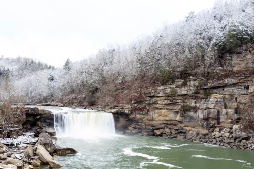 The landscape around Cumberland Falls is whitewashed during a wintery season with icy tree branches, snow-covered rocks, and cold flowing water.