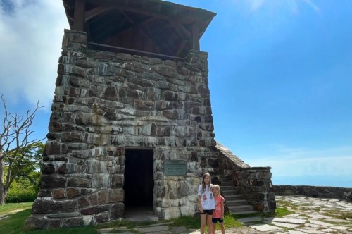 Two little girls stand in front of the tall stone tower with a dark doorway and an inviting staircase leading to the top on the right.
