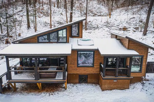 A tanish-brown modern-looking cabin in a wintery wonderland as a fresh layer of snow covers the roof and ground.
