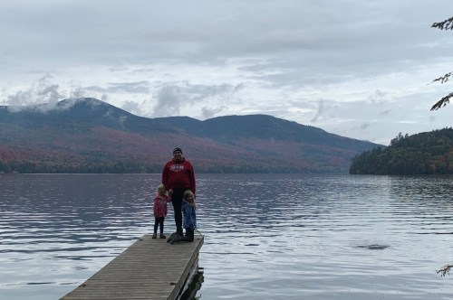 A family standing in front of a large pretty lake with tall and colorful mountains in the background on a crisp fall day.