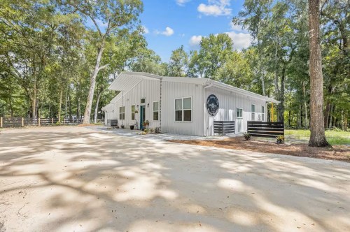 A white barndominion with a large concrete driveway and surrounded by a forest of trees.